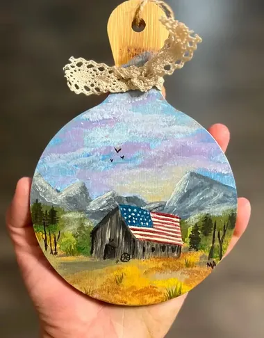 A round wood cutout tied with lace, painted with a barn and an American flag under a mountain sky.