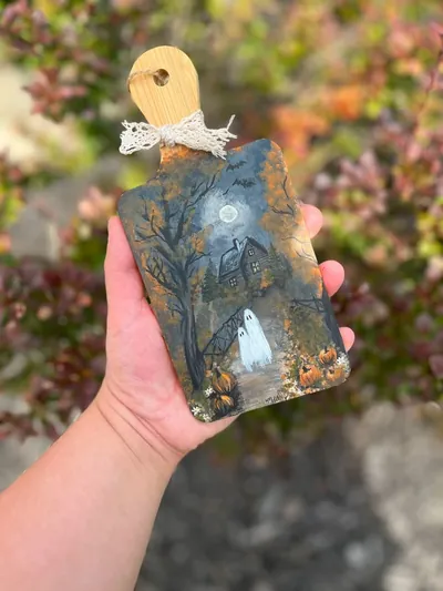 A wooden cutting board held in a hand, painted with ghosts, a haunted house and a harvest moon.