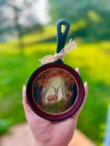 A cast-iron skillet held in a hand, painted inside with a whitetail deer in autumn woods.