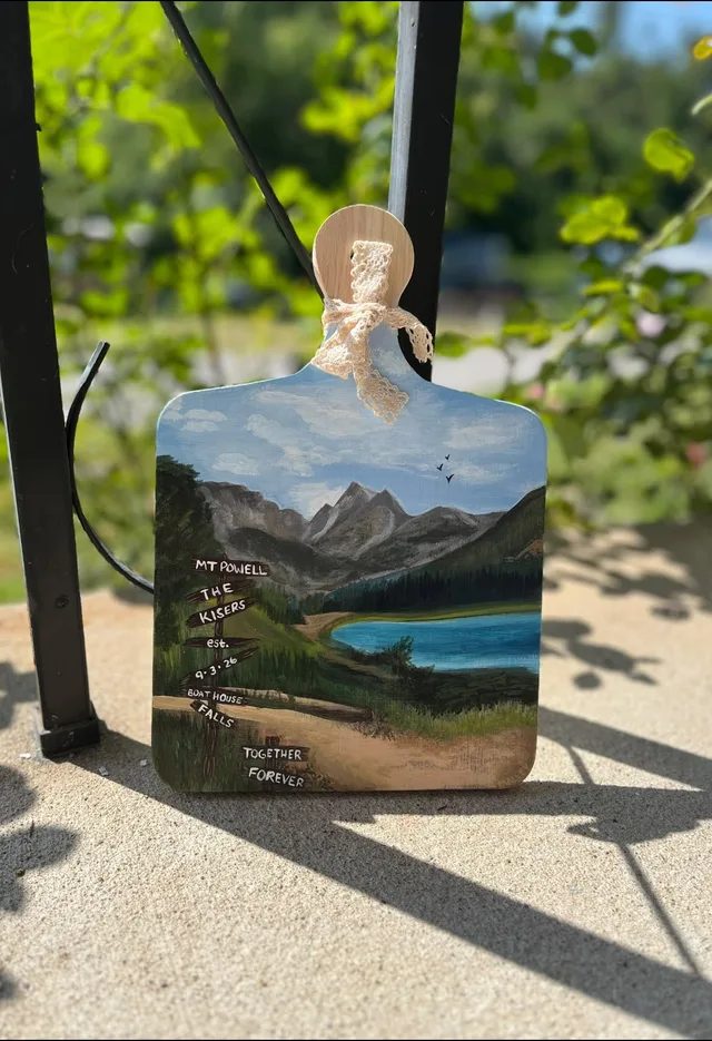 A wooden board painted with a mountain lake, finished with hand lettering.